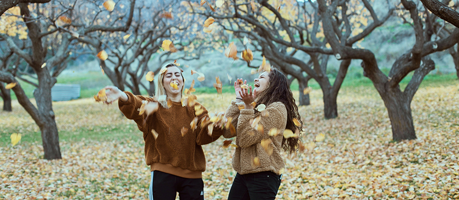 White woman, Hispanic woman in fuzzy jackets throwing leaves into air laughing