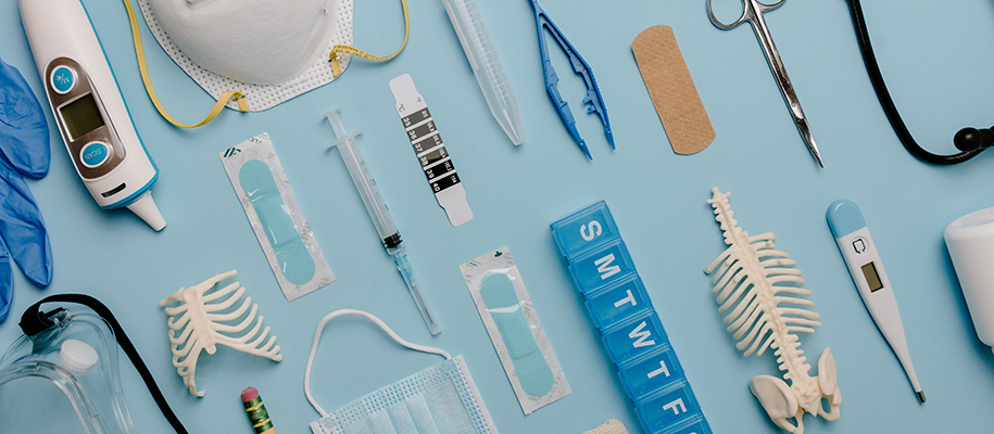 Thermometer, pill case, needle, more medical supplies laid out on blue backdrop