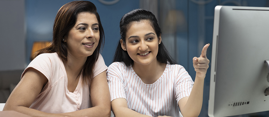 Indian daughter pointing at computer screen with mom, both smiling in T-shirts