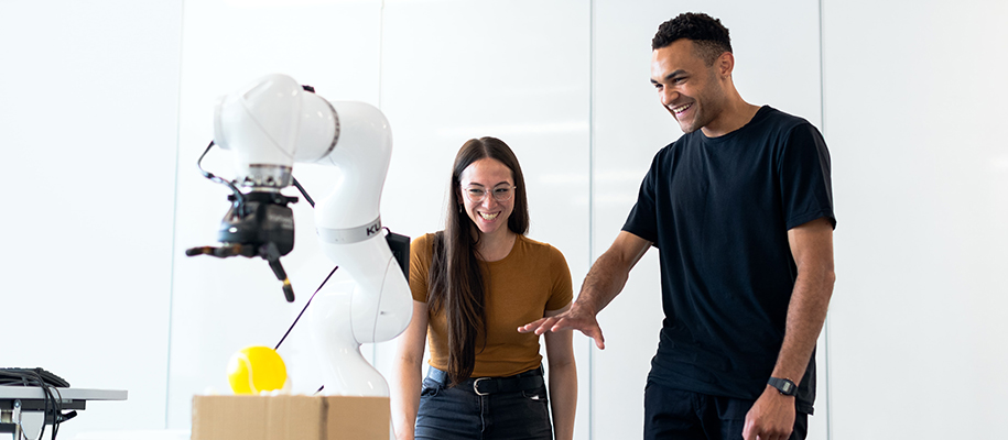 Man and woman smiling and gesturing at robotic arm poised to pick up ball