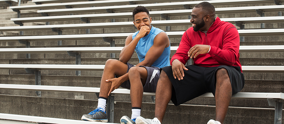 Black coach laughing with young Black man in stands, both in workout gear