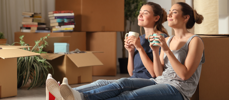 Two White women sitting on floor, surrounded by packed boxes, with coffee cups