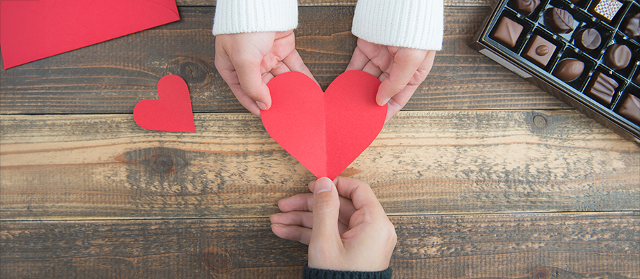 White hands in white sweater handing paper heart to White hand in black sweater