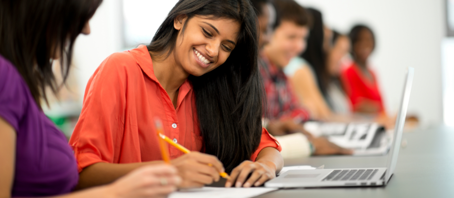 Indian woman in orange shirt smiling, working with another student in classroom