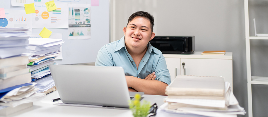 Asian man with down syndrome at office desk, piles of paper and project board