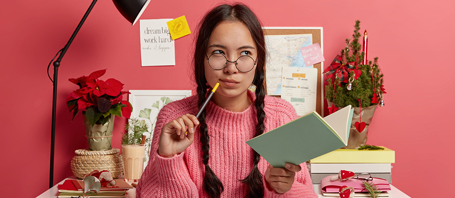 Young Asian woman in pink sweater at festive desk thinking with pencil, notebook