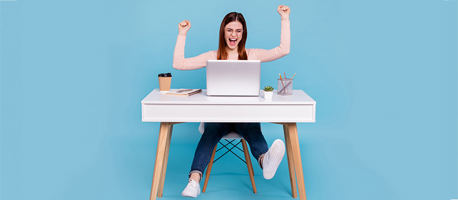 White woman in pink shirt at white desk with laptop, arms up in excitement