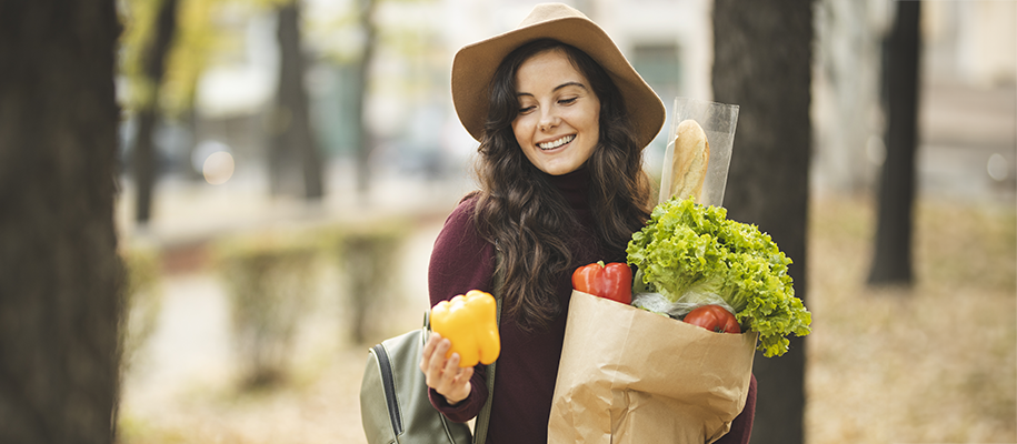 White woman with long dark hair, hat, carrying bag of veggies, smiling at pepper