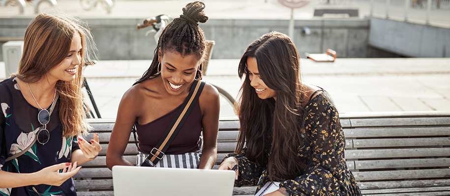 Three racially diverse women in nice clothes on bench with laptop, smiling