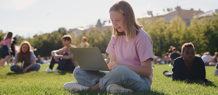 Blonde teen girl in pink shirt on campus lawn with laptop surrounded by others