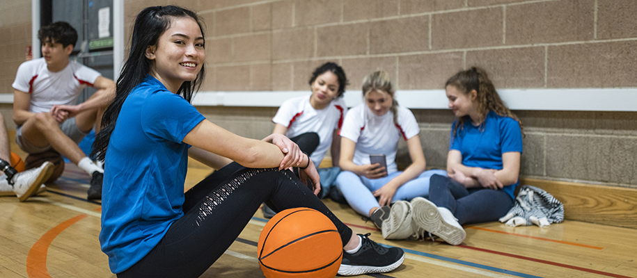 Group of students in workout clothes in gym, focus on Asian girl with basketball