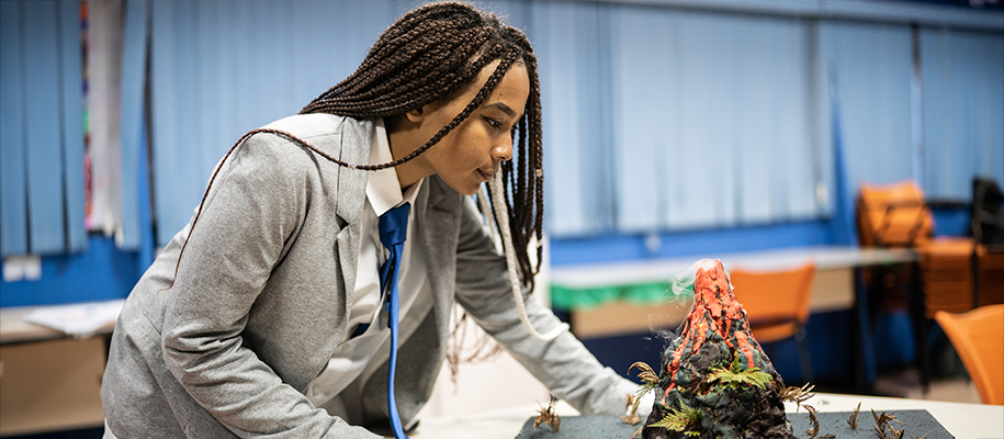 Black girl with braids in blazer and blue tie leaning over science fair volcano