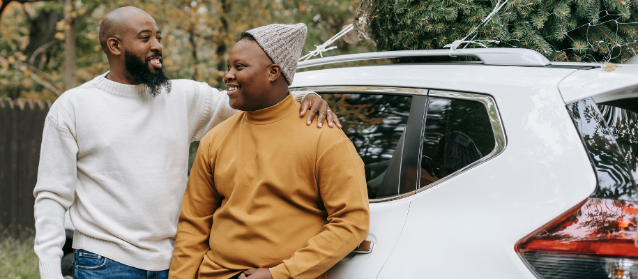 Black father with arm around teen son, leaning against white car with Christmas
