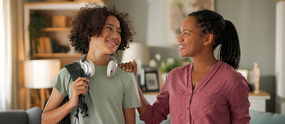 Black teen with backpack and headphones next to mom with hand on his shoulder