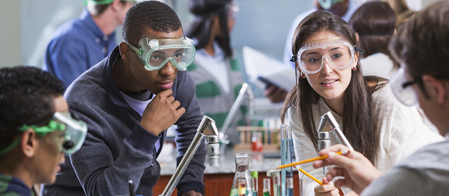 White woman, Black man in science class with lab goggles, listening to teacher
