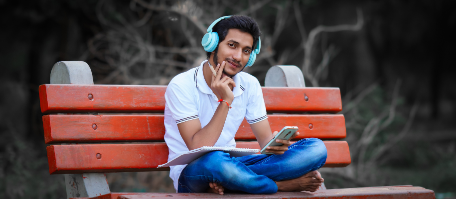 Indian man in white shirt with blue headphones, phone, sitting on bench barefeet