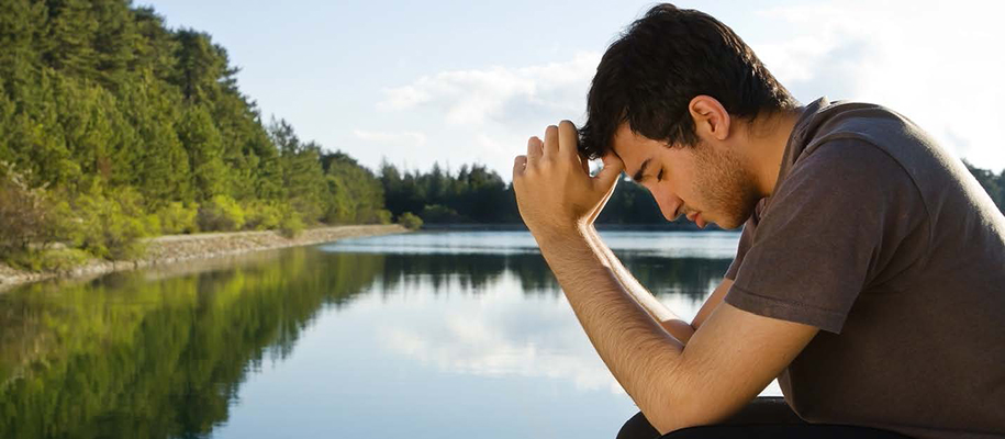 Young White male in gray T-shirt with hands to head, praying by lake
