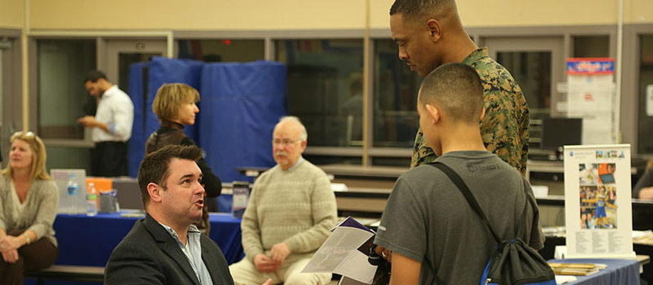 Tall man in fatigues next to shorter man with backpack talking to man in suit