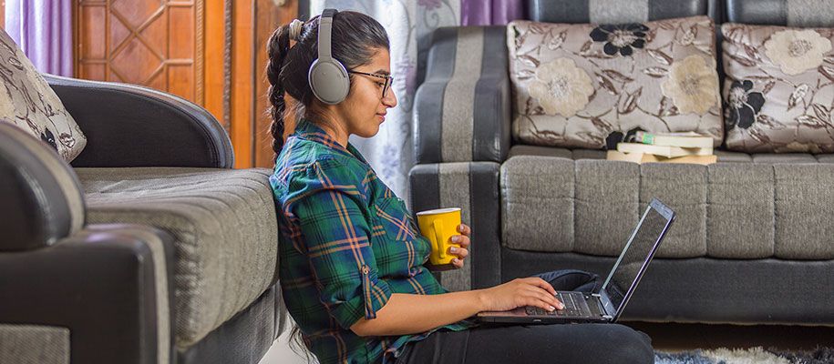Indian female student sitting on floor at home with laptop, headphones, mug