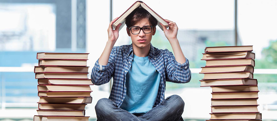 White male student sits between stacks of books, one on head looking concerned
