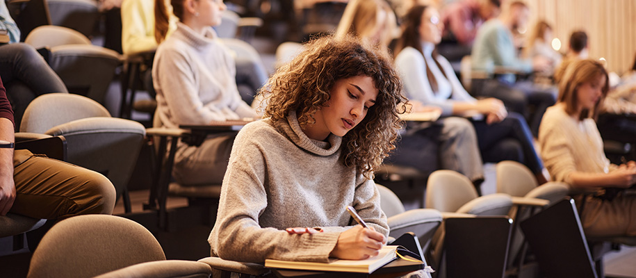 Latina woman in grey sweater writing in notebook in front row of seminar class