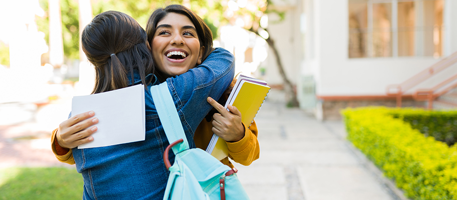 Hispanic girl holding paper, hugging another woman with teal backpack on campus