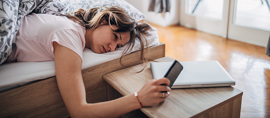 Young White woman lying in bed, hand propped on bedside table with phone, laptop