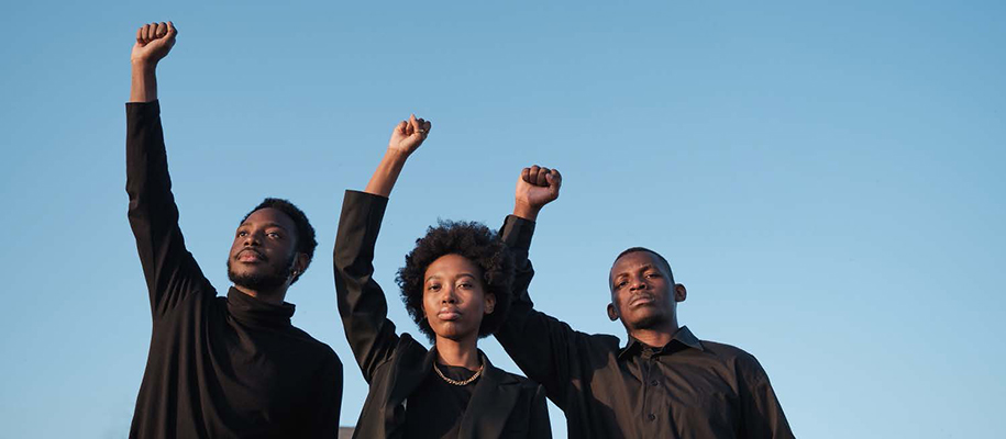 Two Black men and one Black woman in all black clothes outside with raised fists