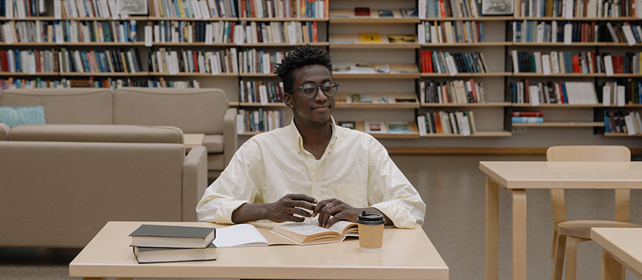 Young Black man in white button-up shirt, glasses at desk in library, with books