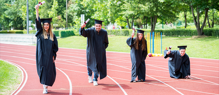 Four students in black graduation caps and gowns hold up degrees on racetrack