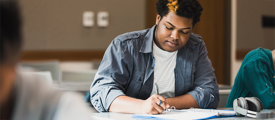 Black male student with yellow stripe in Afro writing notes in class
