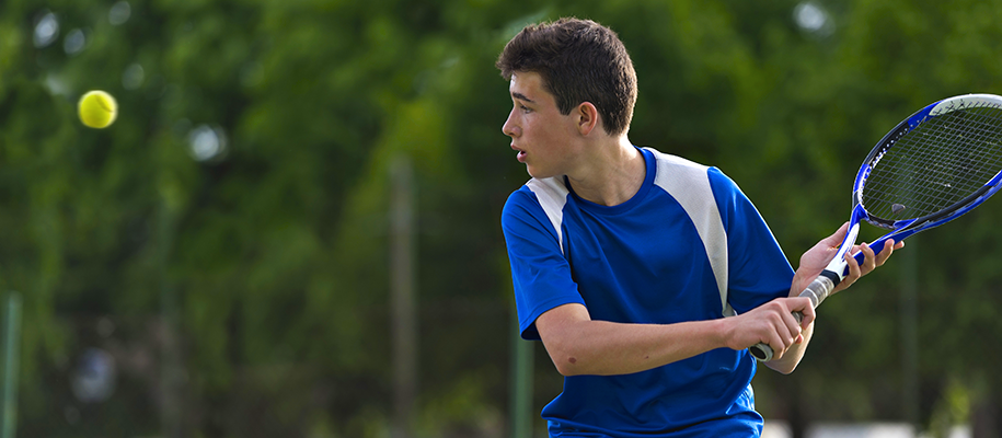 White teen boy in blue tennis uniform with posed racket, eyes on tennis ball