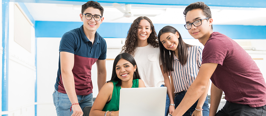 Group of Latino/a teens smiling at camera in classroom, one at desk with laptop