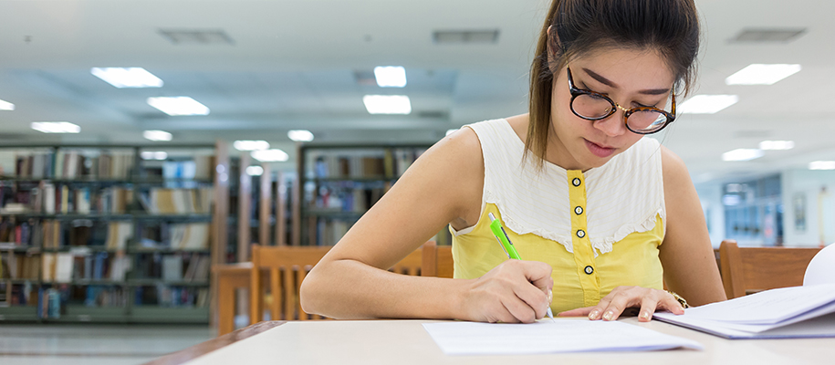 Asian woman in yellow blouse and glasses writing essay in a library