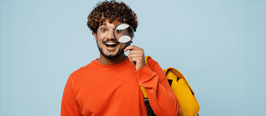 Indian teen in orange shirt with magnifying glass to his eye and yellow backpack