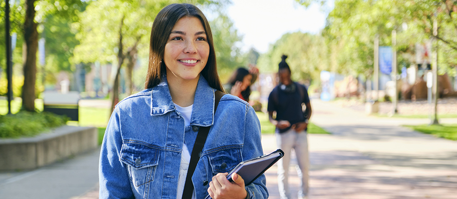 Young woman with long brown hair in denim jacket, smiling on college campus