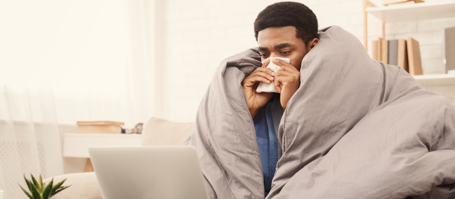 Young Black man wrapped in comforter on couch with tissue to nose and laptop