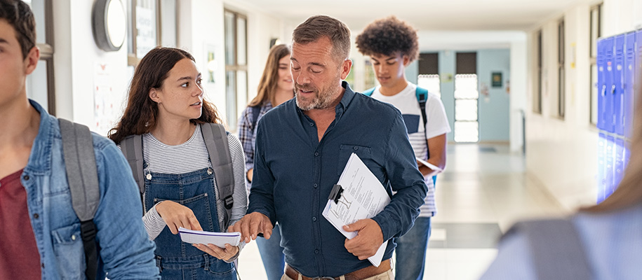 Young woman in overalls, showing notebook to male teacher carrying papers