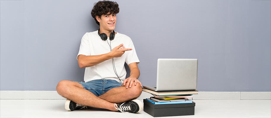 White teen boy in white T-shirt sits, headphones around neck, points at laptop