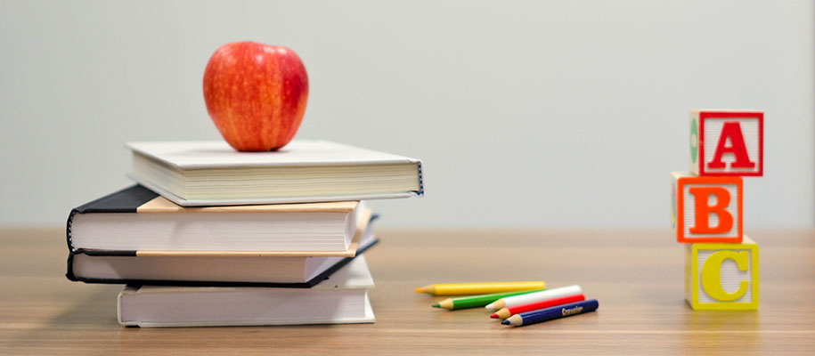 An apple sits atop a stack of books on a desk, next to some lettered blocks