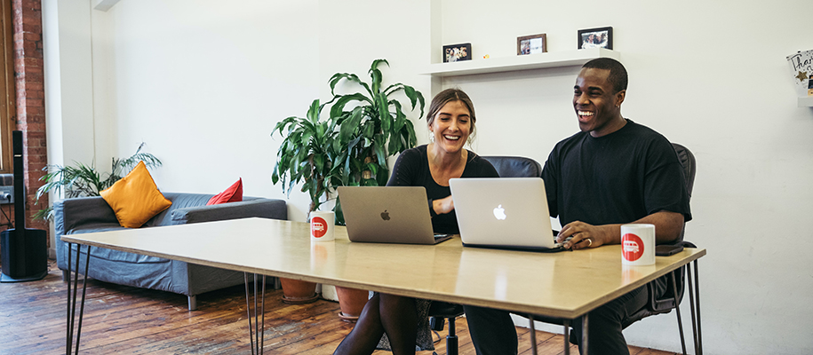 White woman and Black man smiling at a desk together with coffee and laptops