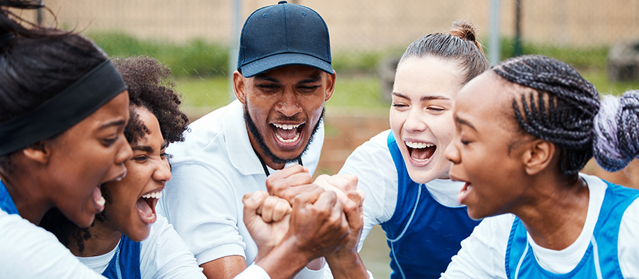 Circle of three Black and one White female athletes cheering with Black coach