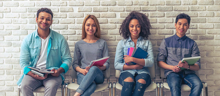 Four diverse students sitting in chairs against white brick wall with books