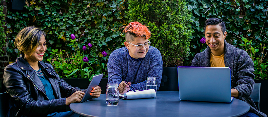 Three people sitting in front of ivy at an outdoor table with laptop and tablet
