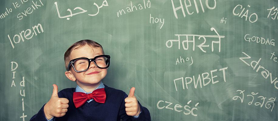 Kid in glasses smiles in front of chalk board with words in various languages