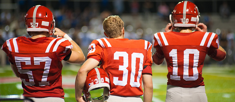 Three football players in red and white uniforms, man in middle with helmet off