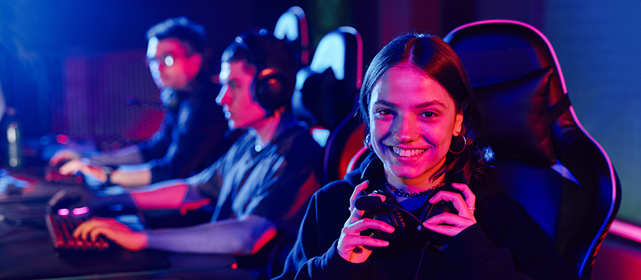 White female gamer with black smiling at camera, two White males playing in back