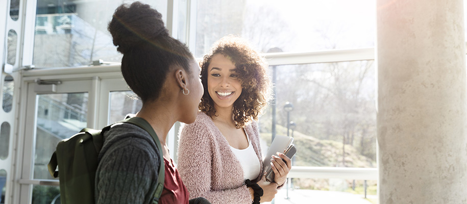 Two black women with backpacks, notebooks smile at each other in campus building