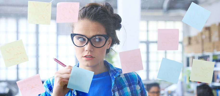 Young White woman wearing glasses, writing on Post-It notes on glass wall
