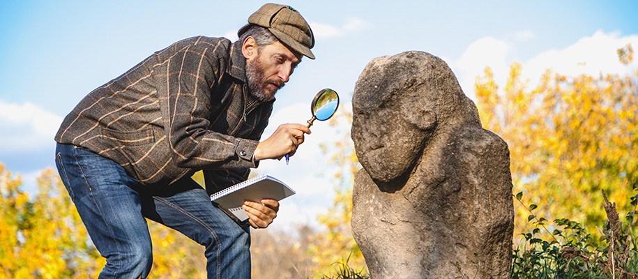 White man with beard in cap with notebook, magnifying glass observing statue